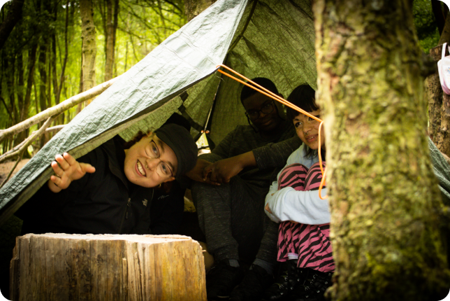 A group of people in a tent in a forest. Two of the people are peaking their heads through the tent entrance.