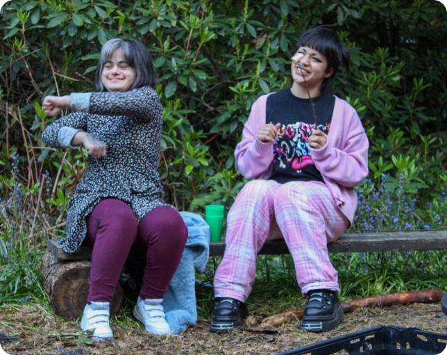 2 people smiling sitting on a log bench in a forest. One is holding a marshmallow on a stick.