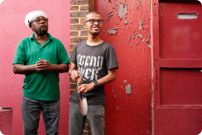 An older person and a young person in front of a red wall. The young person is holding a hand shovel.