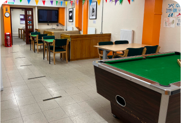 A room with tables, chairs and a pool table. The walls are white and orange and there is colourful bunting near the ceiling.