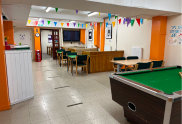 A room with tables, chairs and a pool table. The walls are white and orange with colourful bunting near the ceiling.