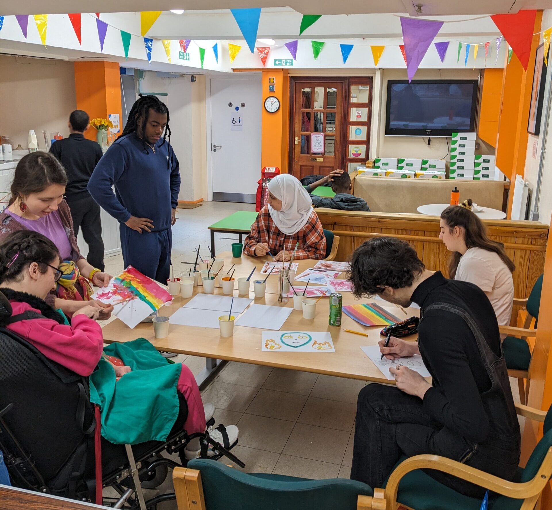 A group of 6 people sitting around a table filled with pieces of paper and pots of paint. Many of them are painting.