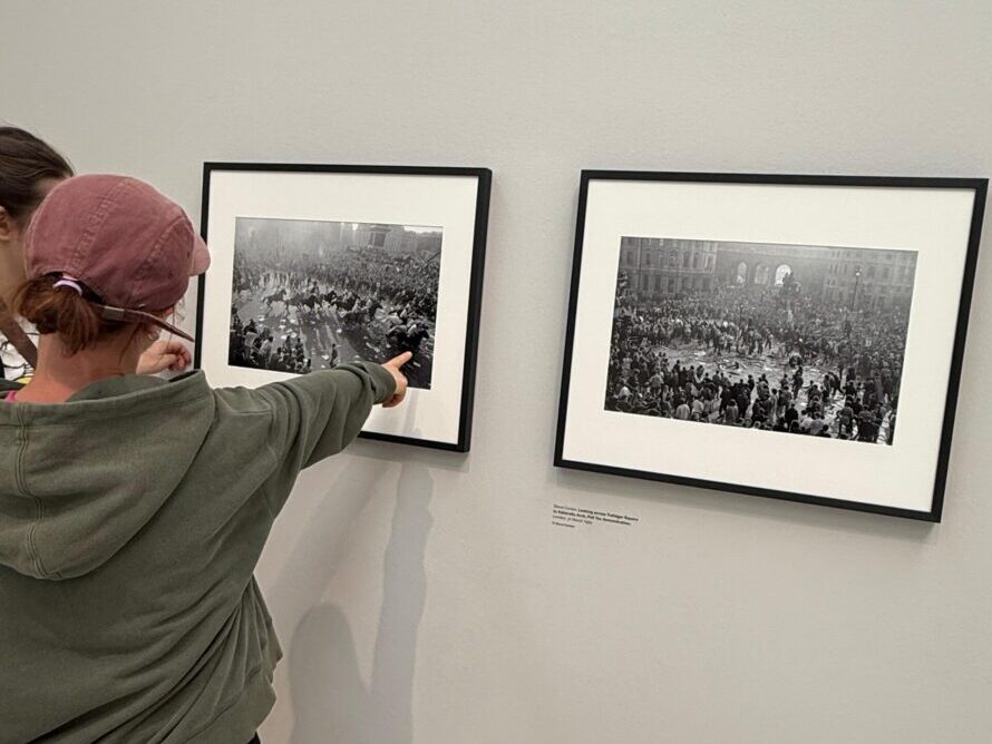 Two people looking at two framed black and white pictures on a wall. One of the people, wearing a green jumper and pink cap is pointing at one of the pictures. The pictures show large groups of people.