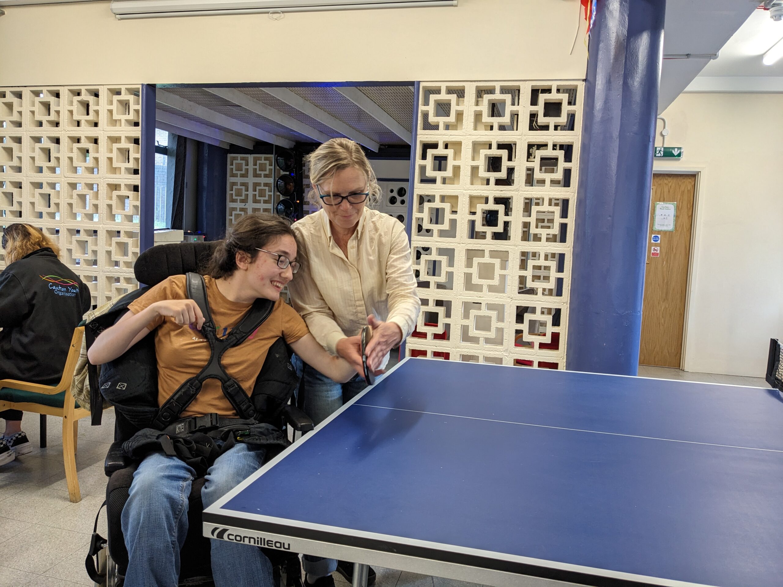 Young person with long dark hair in a wheelchair and an older person playing table tennis.