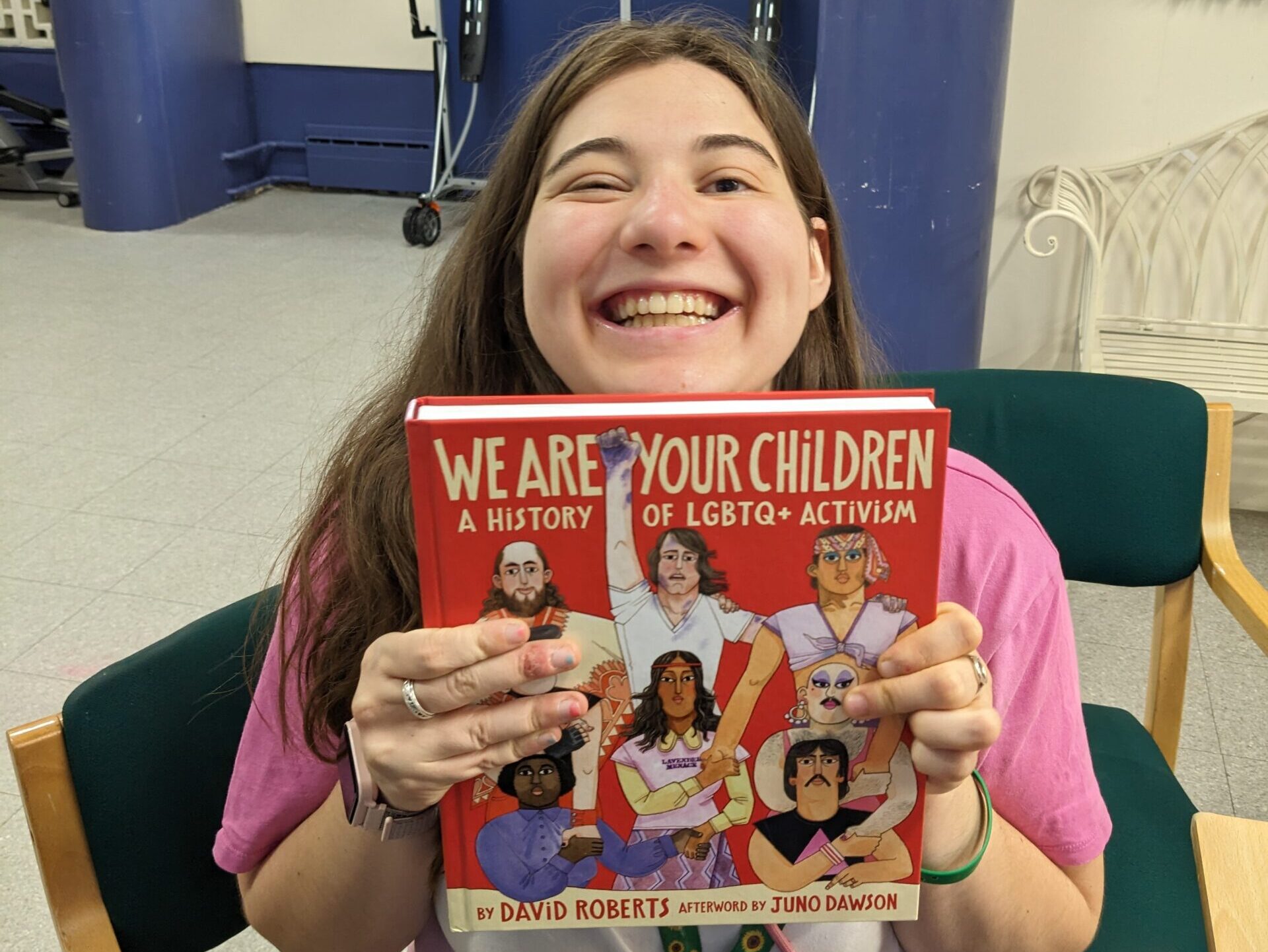 Young person with long dark blonde hair wearing a pink T-shirt smiling and holding up a book titled "We are your children: A history of LGBTQ+ activism".