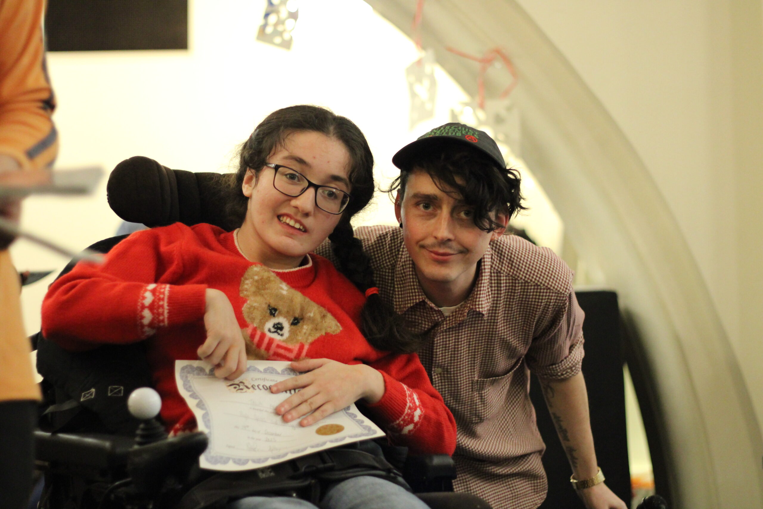 A young woman wearing glasses using a wheelchair and a male youth worker next to her. They are both smiling.
